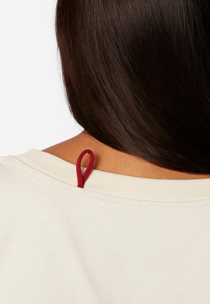 Detailbild einer roten Schlaufe am Pyjama T-Shirt in der Farbe „moon white“ an der Nackennaht.